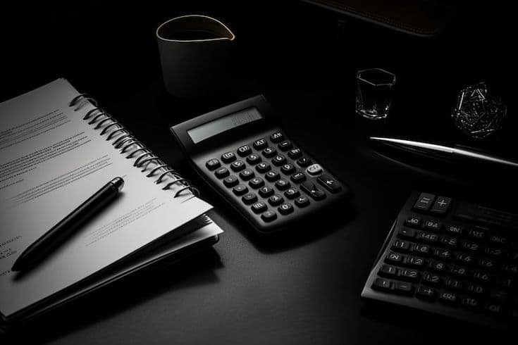 Black calculator, spiral notebook, and pens on a dark desk with dramatic, low-key lighting.