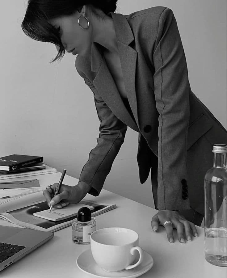 Woman in a blazer leaning over a desk, writing on a notepad near a coffee cup.