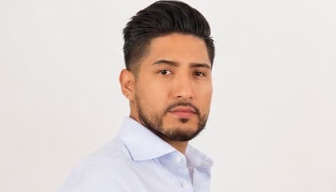 Man with dark styled hair and a beard wearing a light blue pinstriped button-down shirt.